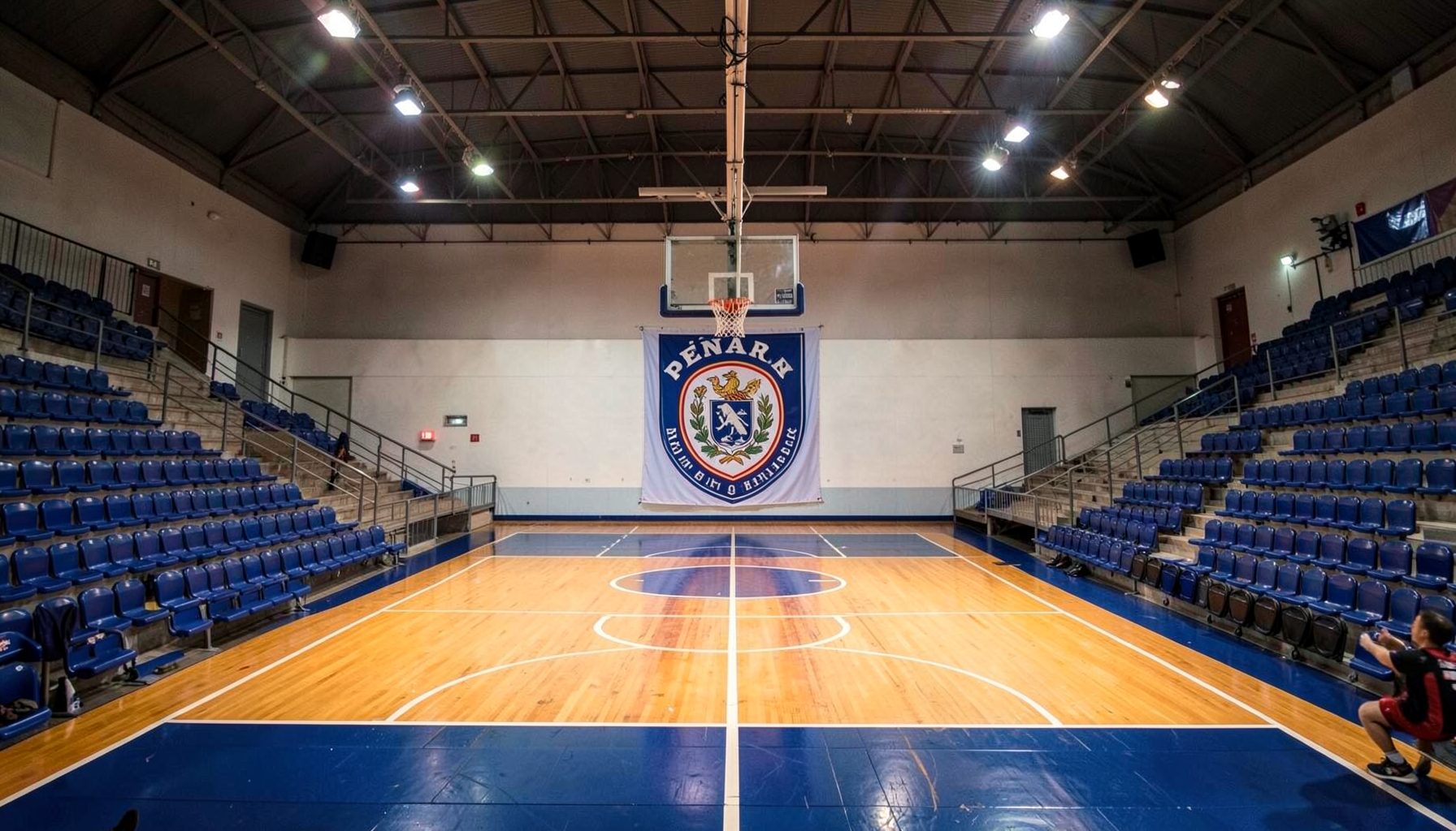 Cancha de básquet con el escudo de Peñarol en el centro, iluminada por reflectores cálidos, rodeada de gradas vacías en tonos azul marino y blanco.