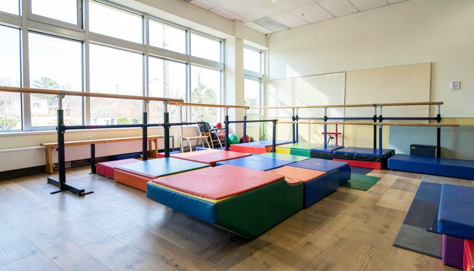Sala de gimnasia con colchonetas coloridas, barras bajas y luz natural entrando por ventanales amplios