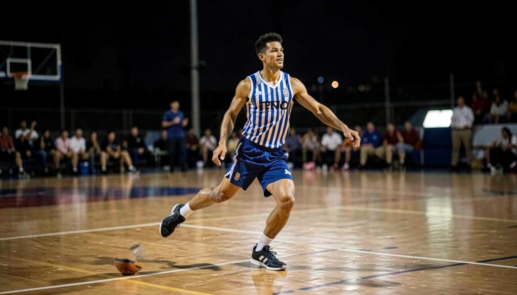 Jugador de básquet en acción sobre cancha de parqué, camiseta a rayas azul y blanca, ambiente de partido nocturno
